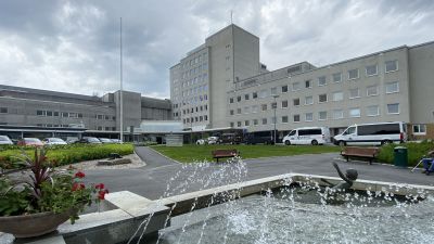 En sjukhusbyggnad. I förgrunden ses en fontän omgiven av ett grönområde.