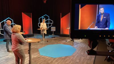 Ett foto taget bakom kullisserna i en tv-studio inför en diskussion. På bilden syns tre personer stå vid varsit bord, en fjärde person syns på en tv-skärm i hörnet.
