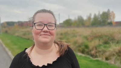 Ann-Sofie Holmberg, en dam med svart skjorta och svartbågade glasögon, står vid en gulnande gräsmatta på hösten.
