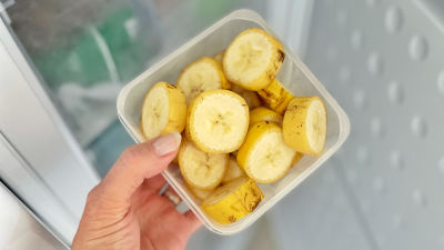 En hand håller i en plastburk fylld av skivade bananer med skalat på. Personen håller burken mot en öppen frys.
