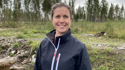 En kvinna med mörkblå jacka och mörkt hår poserar framför skog.