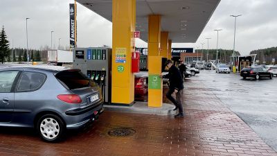 En man i svarta kläder står vid en gul pelare vid en bensinstation för att tanka sin silverfärgade bil.