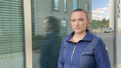 En kvinna står framför ett fönster. Hon ser in i kameran, och ser allvarlig ut.