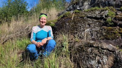 En kvinna med blå-vita utekläder och grönt hårband sitter på en klippa som kantas av gräs och hö.