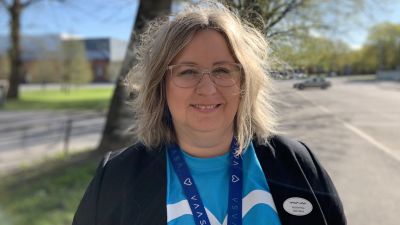 Kvinna står utomhus i vårgrönska, iklädd blus med Vasa stads logo.