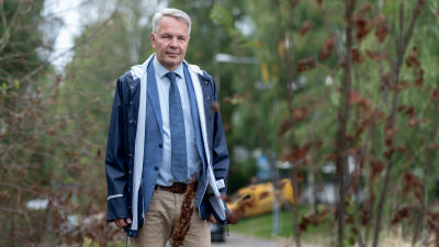 Pekka Haavisto fotograferad utomhus. Han bär en regnrock ovanpå en blå kragskjorta. Han bär slips och beigea byxor.