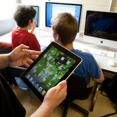 Elever jobbar vid en dator. En lärare står bredvid och håller i en pekplatta.