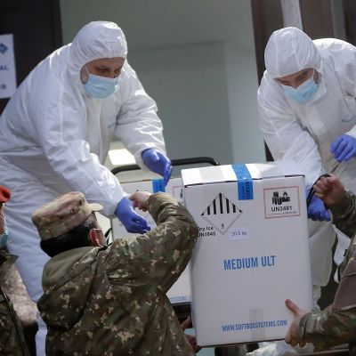 Soldater lyfter upp en låda med coronavaccin åt två personer i skyddsutrustning som tar emot lådan.