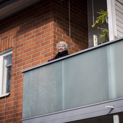 En äldre kvinna står på en balkong och tittar ut. Hon ler.