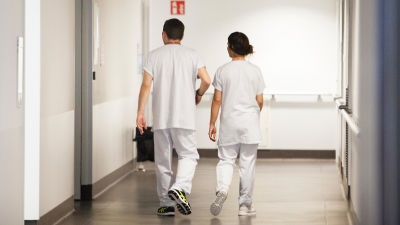 Två sjukskötare klädda i vita uniformskläder går i en sjukhuskorridor. De har ryggarna vända mot kameran.
