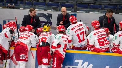 Sportspelare i vita speltröjor står vid sargen och lyssnar på sina tränare.