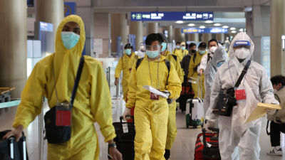 Människor i skyddsutrustning, bland annat munskydd och visir, går genom en flygplats med resväskor i händerna.