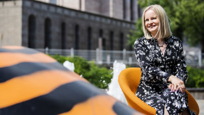 Riikka Purra sitter på en stol utomhus. Hon ler och tittar in i kameran.