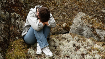 En kvinna sitter med huvudet nedböjt dolt i armarna klädd i grå jacka, blåa jeans och vita fritidsskor på ett stenblock i naturen.