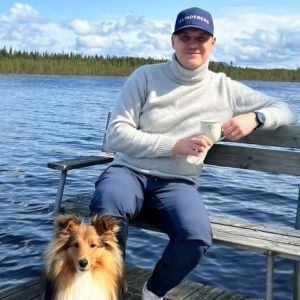 Mies istuu laiturilla, takana vettä. Edessä istuu koira. Miehellä kädessä kahvikuppi. 