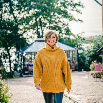 Maria Sundblom Lindberg kommer leende gående mot kameran. Somrig bakgrund med lävträd, växthus och knuten på ett gult trähus.