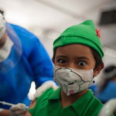 Ett barn i munskydd får coronavaccin av en sjukskötare i skyddsutrustning.