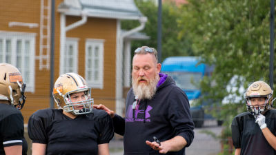 En skäggig man och kvinnor i amerikansk fotboll-spelutrustning är på en gräsplan.