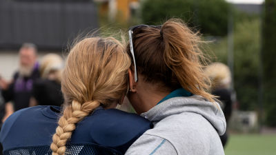 Ryggen och bakhuvudet av två kvinnor som kramas, i bakgrunden idrottsplan.
