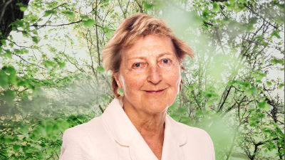 Ulla Tillander-Godenhielm småler in i kameran framför en grönskande bakgrund.
