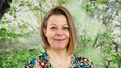 Anette Rönnlund-Nygård småler in i kameran framför en grönskande bakgrund.