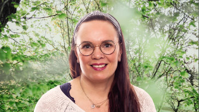 Anna Kolster-Weckström småler in i kameran framför en grönskande bakgrund.