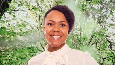 Elina Sagne-Ollikainen småler in i kameran framför en grönskande bakgrund.