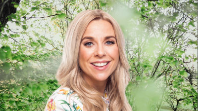 Krista Siegfrids småler in i kameran framför en grönskande bakgrund.