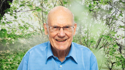 Per-Erik Lönnfors småler in i kameran framför en grönskande bakgrund.