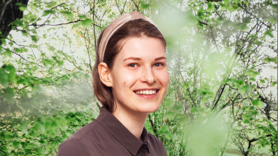 Sara Jakobsén småler in i kameran framför en grönskande bakgrund.