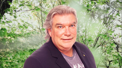 Mikael Lindfelt småler in i kameran framför en grönskande bakgrund.