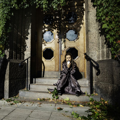 På bilden syns författaren och regissören Caroline Ringskog Ferrada-Noli. Hon sitter på en trappa iklädd en svart jacka och håller i en take away-kaffe. 