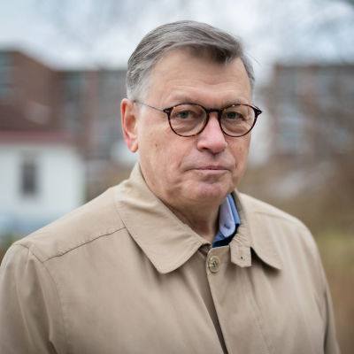 En man i grått hår och glasögon står utomhus. Han har på sig en beige jacka och tittar in i kameran.