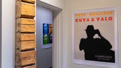 En vit vägg med en tavla där det står Pave Maijanen och en bit av stockar.
