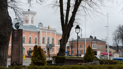 Parkträd i förgrunden, bakom dem en stad med hus och torg.