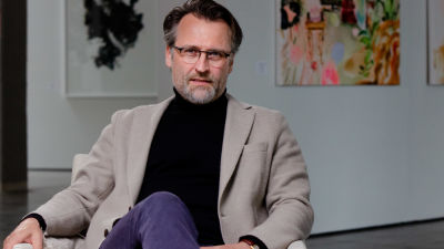 Jan Söderblom sitter med benen i kors i en beige fåtölj. Färggranna tavlor hänger på väggarna i bakgrunden.