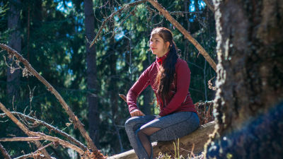 En kvinna iklädd sportkläder sitter på en sten ute i skogen. Hon ser nöjd ut. Solen lyser på hennes ansikte.
