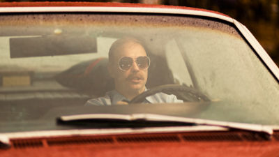 Auton tuulilasin läpi otetussa kuvassa näkyy viiksekäs mies (näyttelijä Jarkko Lahti) auton ratissa aurinkolasit päässään.