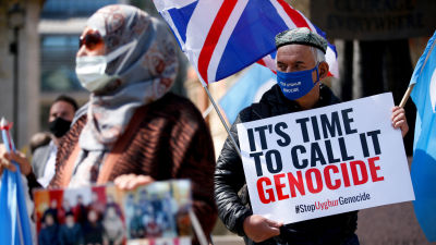 En man i blått munskydd håller upp ett plakat med texten "It's time to call it genocide".