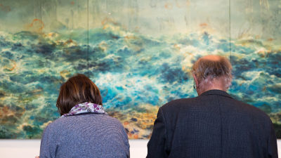 En kvinna och en man sitter på en bänk framför ett konstverk på ett museum. 