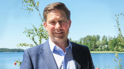 Thomas Blomqvist klädd i kostym står vid havet i Bromarv skärgårdshamn. I bakgrunden syns hav och träd. Han ler och tittar in i kameran.