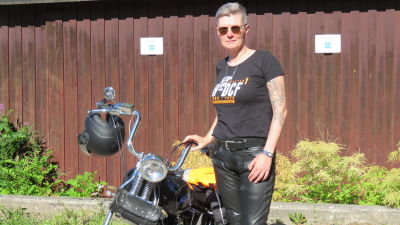 En kvinna med kort hår, solglasögon, svart t-shirt och läderbyxor står vid en motorcykel. Det är sommar och sol.