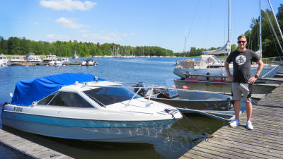 En ganska ung man står på en brygga bredvid sin motorbåt. I bakgrunden både motorbåtar och segelbåtar. Sommar och soligt.