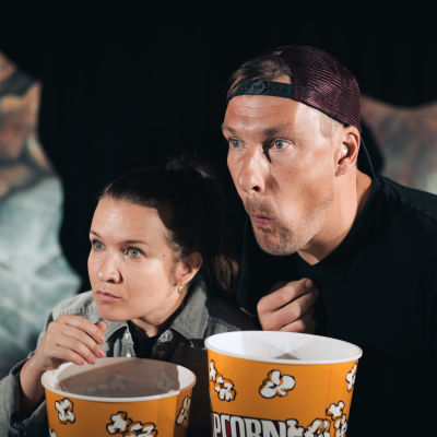 En man och en kvinna med popcornaskar i händerna följer spänt något med blicken.