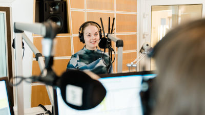 A journalist with a headset on is standing in front of a microphone in a radio studio.