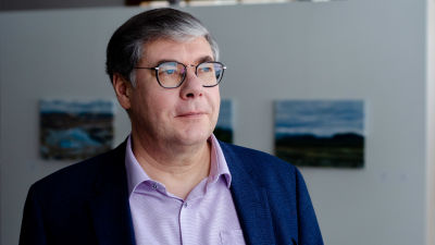 Asko Järvinen ser snett förbi kameran. Bakom honom syns en vit vägg med tre små tavlor.