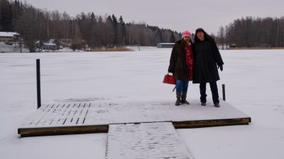Två kvinnor står på en snöig brygga. Bakom dem syns Veckjärvi sjö täckt av is. På andra sidan sjön syns några blå byggnader vid Hasselholmens badstrand.