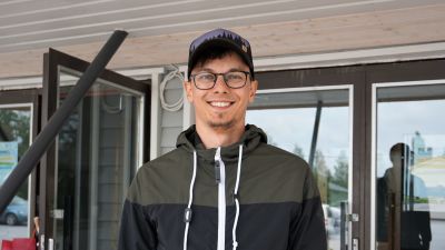 Ung man i keps och glasögon står framför en vit byggnad.