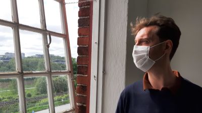 André Peterdi, en man med vitt munskydd och blå tröja, står vid ett fönster med många små rutor och tittar ut på ett sommargrönt landskap.