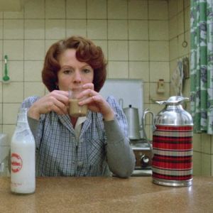 Nainen istuu keittiön pöydän ääressä ja juo kahvia, pöydällä maitopullo ja termoskannu.